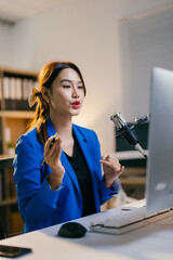 Asian businesswoman is recording a podcast in her home office, speaking into a professional...