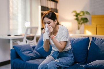 Sick woman on her sofa at home, blowing her nose, surrounded by tissues, medicine, and a warm...