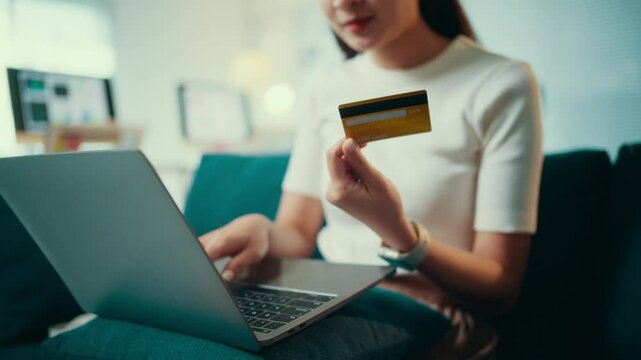 Woman sitting on a sofa using a laptop computer for online shopping. She is holding a credit card and entering the number to complete a purchase