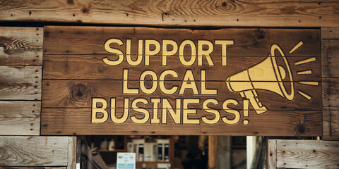 Wooden "Support Local Business!" sign with megaphone graphic hanging on a shop wall. Simple, natural design emphasizes local community support and the importance of small businesses.

