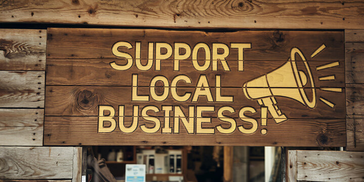 Wooden "Support Local Business!" sign with megaphone graphic hanging on a shop wall. Simple, natural design emphasizes local community support and the importance of small businesses.

