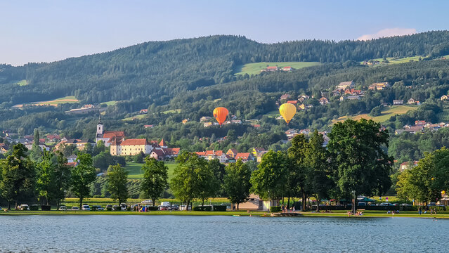 Two colorful hot air balloons float gracefully above bathing lake Stubenbergsee surrounded by rolling hills of East Styrian Hill Country in Styria, Austria, Europe. Travel destination in summer