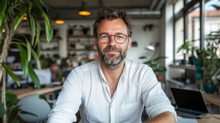 Obraz premium Smiling Professional Man in White Shirt Working in a Bright, Plant-Filled Office, Surrounded by Natural Light and Modern Workspace, Reflecting a Positive and Productive Work Environment