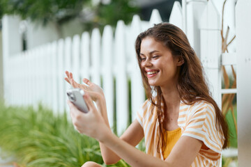 Smiling woman using smartphone outdoors near a white picket fence, showing joy and connection,...