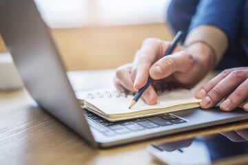 A man's hand writing in a notepad on a laptop, captured in close-up detail, set against a blurred surrounding