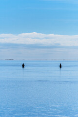 Two fishermen standing waist-deep in the calm Mediterranean Sea under a clear blue sky and Trabucador beach on the horizon with people strolling.