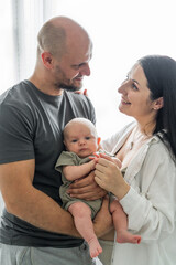 Happy man and woman holding newborn cute baby in arms. Touching look at baby. Atmosphere warmth, family love. Baby quietly sleeping in arms of parents.