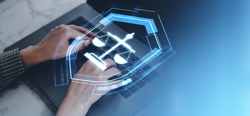 Hands typing on laptop with justice scale icon hologram overlay on blue background.
