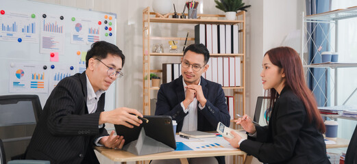Strategic Business Meeting: Focused Asian business professionals collaborate around a table, reviewing data on a tablet and documents.