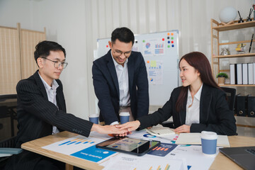 Teamwork Makes the Dream Work: Three business professionals collaborate, their hands stacked in unity over a tablet showcasing data, symbolizing synergy and shared success in a modern office setting.