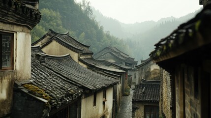 Obraz premium Narrow, cobblestone street lined with traditional Chinese houses with black tiled roofs, nestled in a misty, green mountain landscape.
