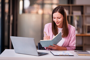 Beautiful professional woman sitting at table smiling while reading important documents with financial graphs and accounting of company.