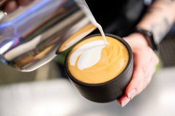 Close-up of hands pouring milk into a black cup of coffee, creating intricate latte art. The scene captures the delicate process of making a perfect latte