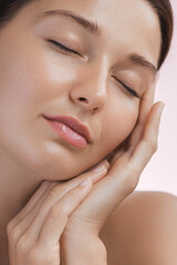 Close-up of a serene young woman with flawless, smooth skin, eyes closed, gently touching her face....