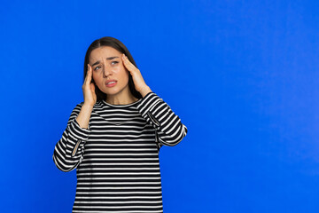 Fototapeta premium Exhausted young woman girl rubbing temples to cure headache problem, suffering from tension, migraine stress, grimacing in pain, high blood pressure isolated alone on blue wall background. Copy-space