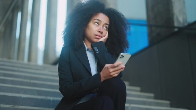 Boring young african american businesswoman scrolling feed of social networks products in internet store on smartphone outdoors Tired exhausted girl without enthusiasm at business centre stairs	