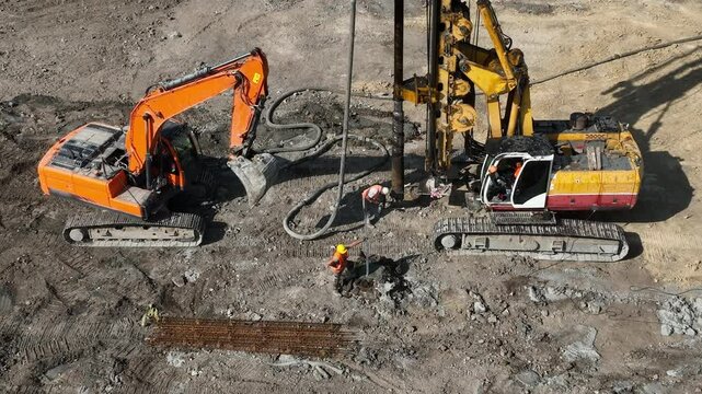 Construction workers operating heavy equipment at a job site. An aerial view of a construction site with workers operating large excavators and drilling machinery.