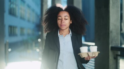 Successful young African American businesswoman hurry to work with takeaway coffee holder for her colleagues walking near modern business buildings outdoors Business morning concept