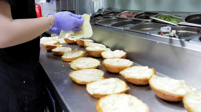 A cook in a fast food restaurant kitchen prepares a cheeseburger and hamburger, placing cheese on the buns. Ideal for scenes about fast food cooking and burger preparation.
