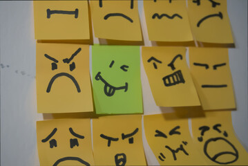 A collection of hand-drawn emoticons on sticky notes, with most showing angry or serious expressions, except for one bright green note featuring a playful, tongue-out smiley, standing out in the group