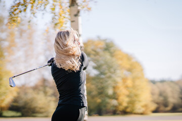 A woman finishing her golf swing outdoors on a sunny day, with flowing hair and autumn trees in the...