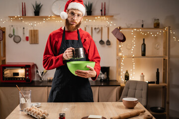 Portrait of man wearing santa hat sifting flour in kitchen decorated for christmas, preparing to bake gingerbread cookies