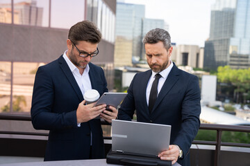 Two businessman on the street, deeply engrossed, eyes fixed on their laptop screen. Two handsome young businessmen in classic suits using laptop. Business men team using laptop outdoor. Business man