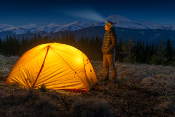 Man mountaineer with headlamp standing near his tent against a backdrop of snowy mountains.