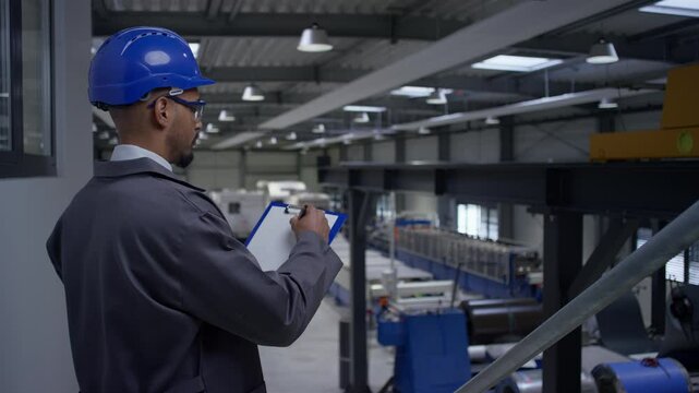 African American man with a white helmet, a manager monitoring productivity in a factory and writing down data, side view. Manufacturing career concept.