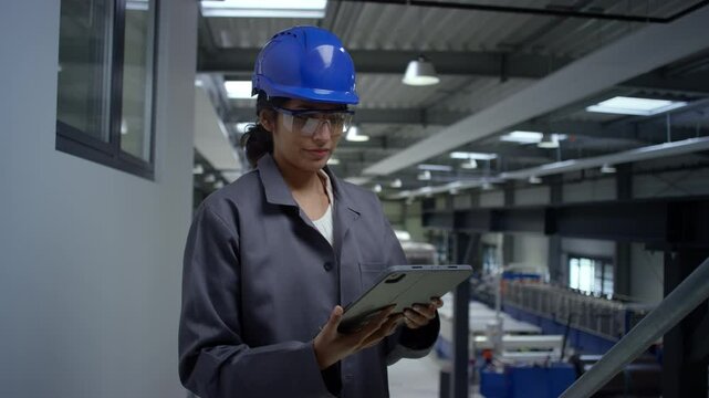 Young woman engineer wearing a blue helmet and work coat posing at a factory holding a tablet, portrait shot. Female professionals in industry concept.