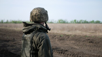 A soldier in camouflage gear stands in an open field, gazing towards the horizon under a clear blue sky. His tactical posture reflects readiness and duty in a rural setting.