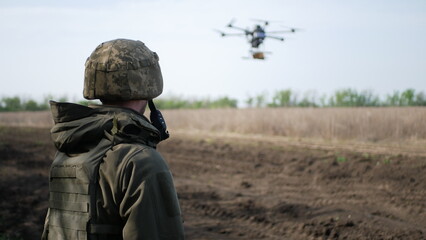 A soldier in camouflage observes a drone hovering above an open field. The drone represents modern...