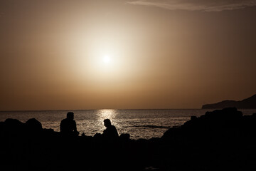 Two people relaxing on rocks by the sea at sunset