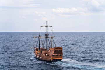 A wooden sailing ship moving through a calm sea under a partly cloudy sky. The ship appears to be...