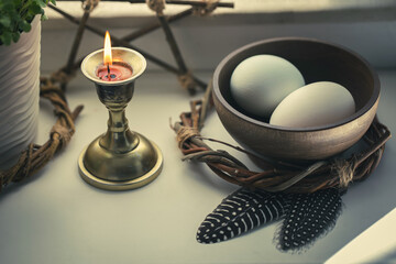 Home pagan set on windowsill with bowl with eggs, pentacle, burning candle and birds feathers, close-up, selected focus.