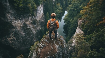 Obraz premium A lone hiker stands at the edge of a cliff, overlooking a serene river surrounded by lush, green trees, embodying adventure and nature's beauty.