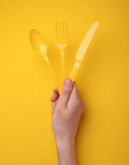 Hand holding plastic yellow fork, spoon and knife on yellow background