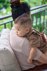 Mother is playing with her 9-month-old son in the summer, in a cool, green setting.
The mother and daughter wore coffee brown clothes. Both are Asian.