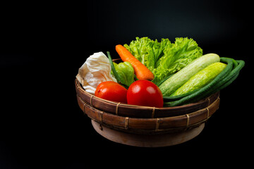 arrangement of vegetables commonly used in indonesian dishes, including tomatoes, carrot, cucumbers, long beans, pumpkin, cabbage, and lettuce. Put on traditional bamboo basket, solid black background