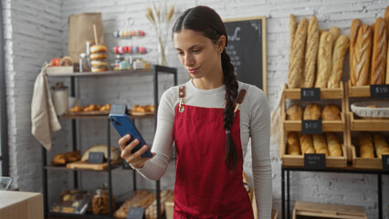 Woman brunette wearing red apron browsing phone in cozy bakery surrounded by fresh breads
