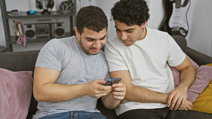 Two men sitting closely on a couch, engaged in looking at a smartphone in a cozy living room.