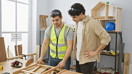Two men in a workshop examine a wooden furniture piece with tools and safety gear around them.