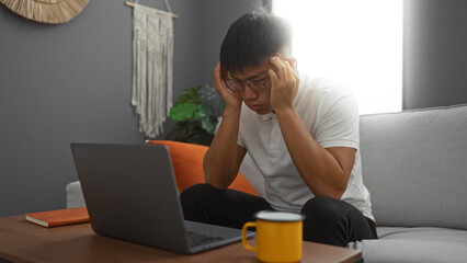 Young man working at home in a living room, appearing stressed while looking at his laptop, surrounded by a cozy interior with a book, coffee cup, and decorative elements.