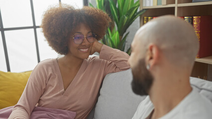 Fototapeta premium Interracial couple smiling lovingly at each other indoors, with a woman and a bald man on a couch, suggesting a comfortable, affectionate relationship.