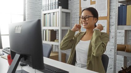 Chinese woman celebrating success at her computer in an indoor office setting, showcasing a professional and joyful moment.