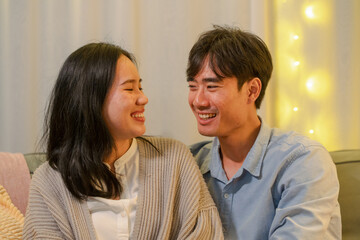 Happy married Asian couple hugging, sitting on cozy couch together, overjoyed laughing woman and man having fun, enjoying leisure time, relaxing on sofa in living room at home, good relationship 