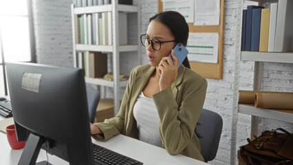 Young, asian, woman talking on phone at office desk with computer in indoor, workplace setting wearing glasses and green blazer in modern interior room, focused on work.