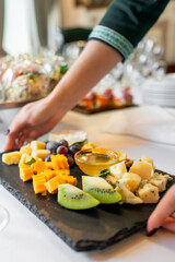 A hand holding a slate tray with an assortment of cheeses and sliced fruits, with wine glasses in the background. Perfect for gourmet dining, catering, or food and beverage themes.