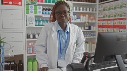 A young african american female pharmacist stands confidently in a modern pharmacy interior behind the counter.