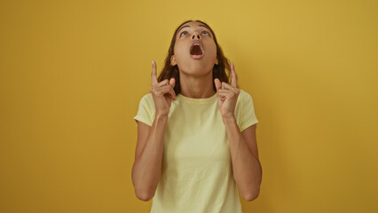 Fototapeta premium Young woman with brunette hair looking upwards in surprise, pointing with fingers, over an isolated yellow wall background.